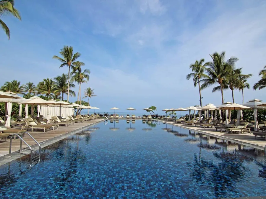 Resort pool lined with palms and cabanas looking out to the Kona coastline at Hualalai Resort