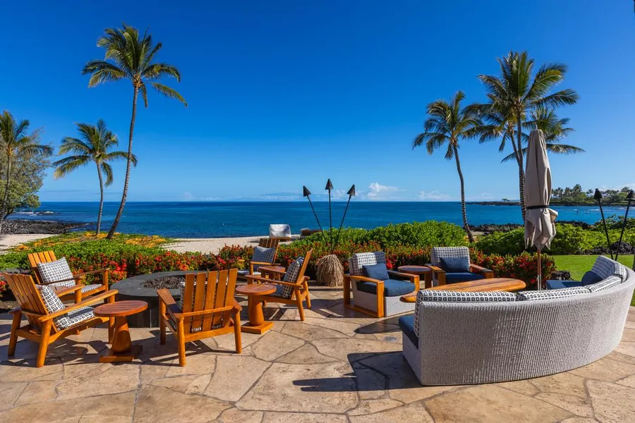 Stone terrace with lounge seating and torches looking through palms to the beach at Kukio