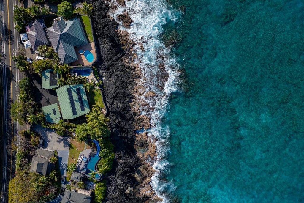 Aerial view of luxury oceanfront homes along the coastline in Kona, Hawaii, showcasing pools and rocky shoreline.