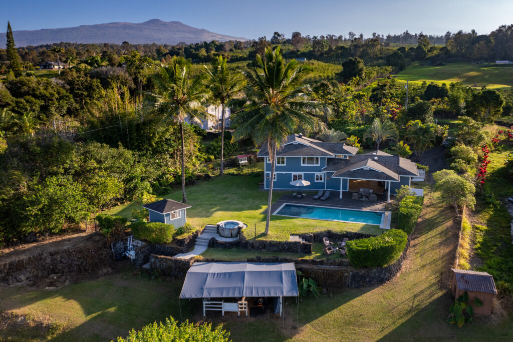 The breathtaking exterior of 76-5876 Hookahi Rd, set against the backdrop of Hualālai volcano, featuring a blue luxury home, tropical landscaping, and a private pool.