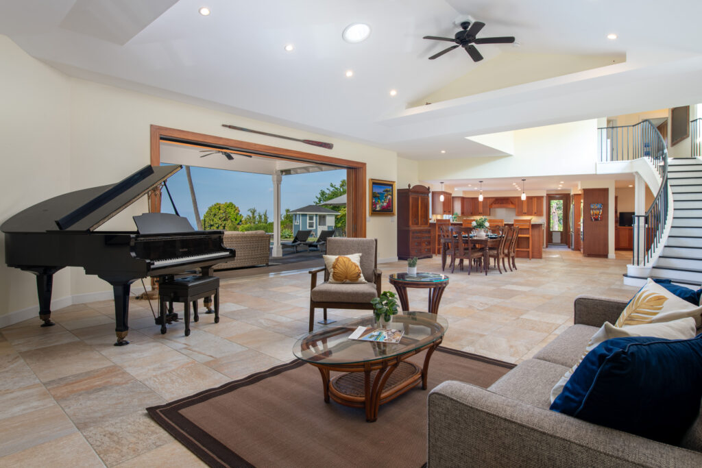 The elegant living room of 76-5876 Hookahi Rd, featuring an open floor plan, a grand piano, modern furnishings, and seamless indoor-outdoor living with panoramic views.